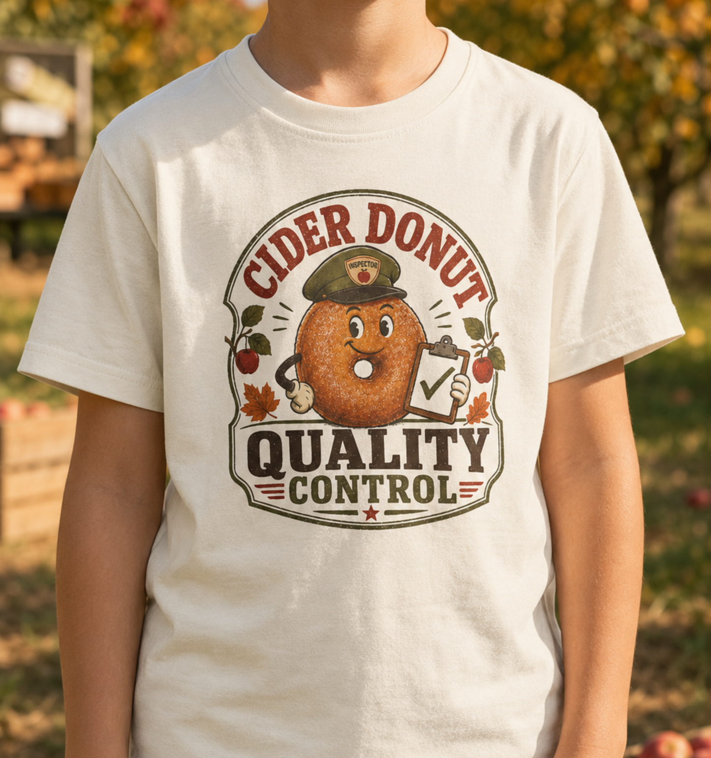 Child wearing a natural youth T-shirt featuring a vintage cider donut inspector graphic, apples, autumn leaves, and the phrase “Cider Donut Quality Control.”