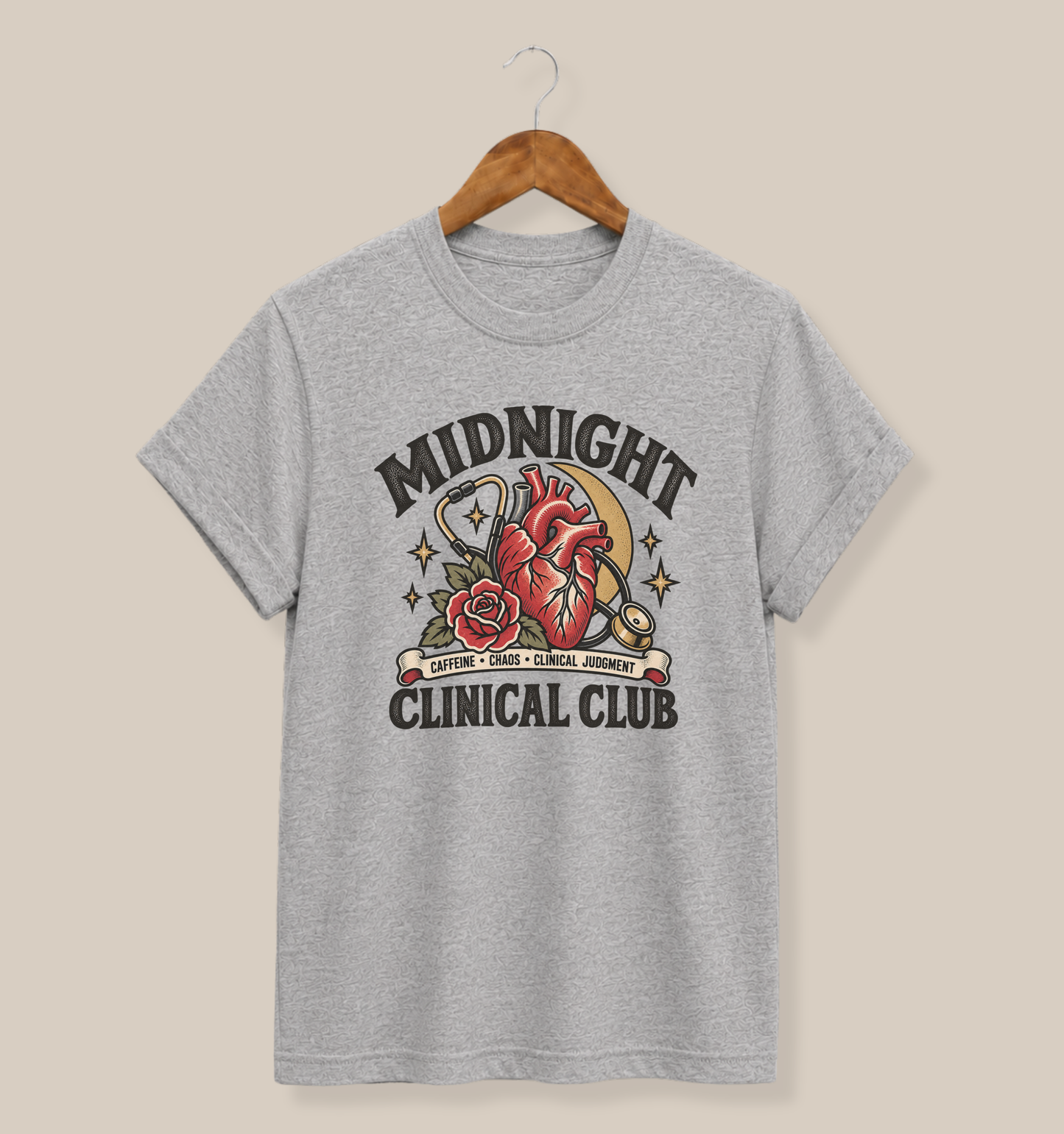 A heather gray short-sleeve t-shirt hanging on a wooden hanger against a solid beige background. The shirt features a detailed graphic on the front with the text 'MIDNIGHT CLINICAL CLUB' surrounding an anatomical heart illustration with a red rose and a crescent moon.