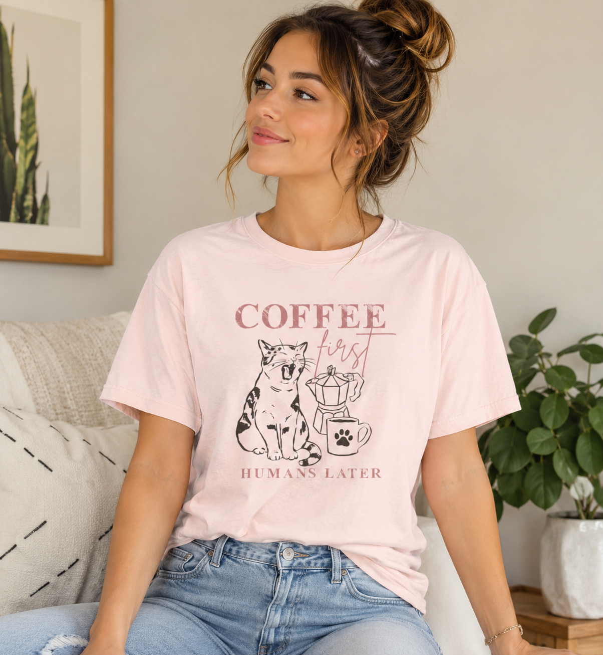 Woman wearing a soft pink Bella + Canvas 3001 t-shirt featuring a “Coffee First Humans Later” graphic with a cat, moka pot, and coffee mug design in a cozy home setting.