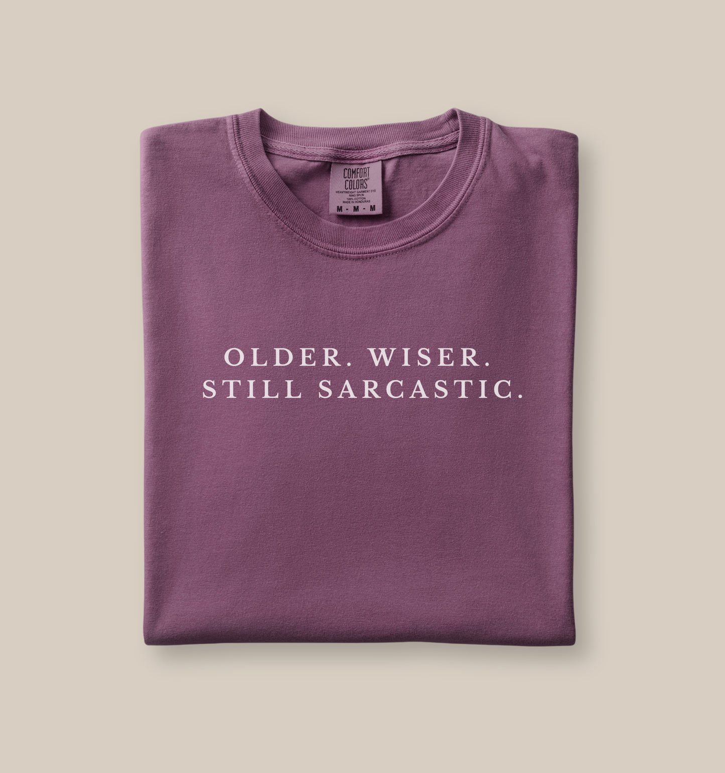 Folded berry t-shirt with text 'OLDER. WISER. STILL SARCASTIC.' on a beige background