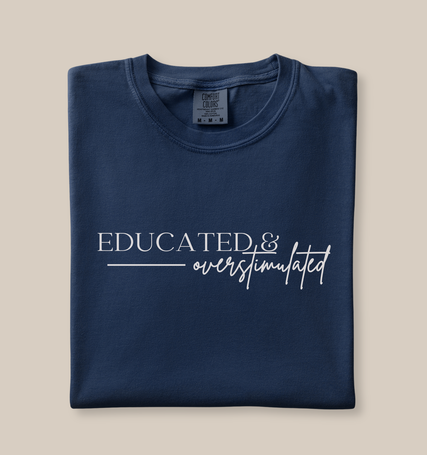 Folded Comfort Colors 1717 t-shirt in navy featuring a minimalist white “Educated & overstimulated” graphic design with mixed serif and script typography.