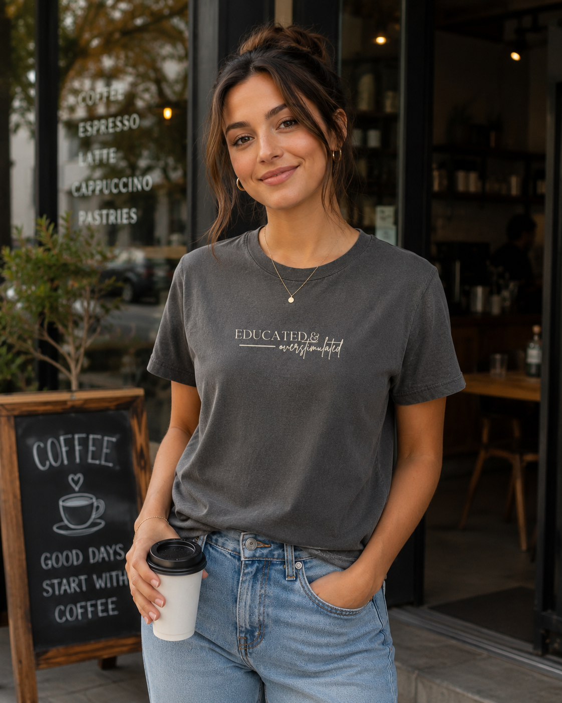 Woman wearing a Comfort Colors 1717 t-shirt in pepper featuring a minimalist “Educated & overstimulated” graphic while holding a coffee cup outside a coffee shop.