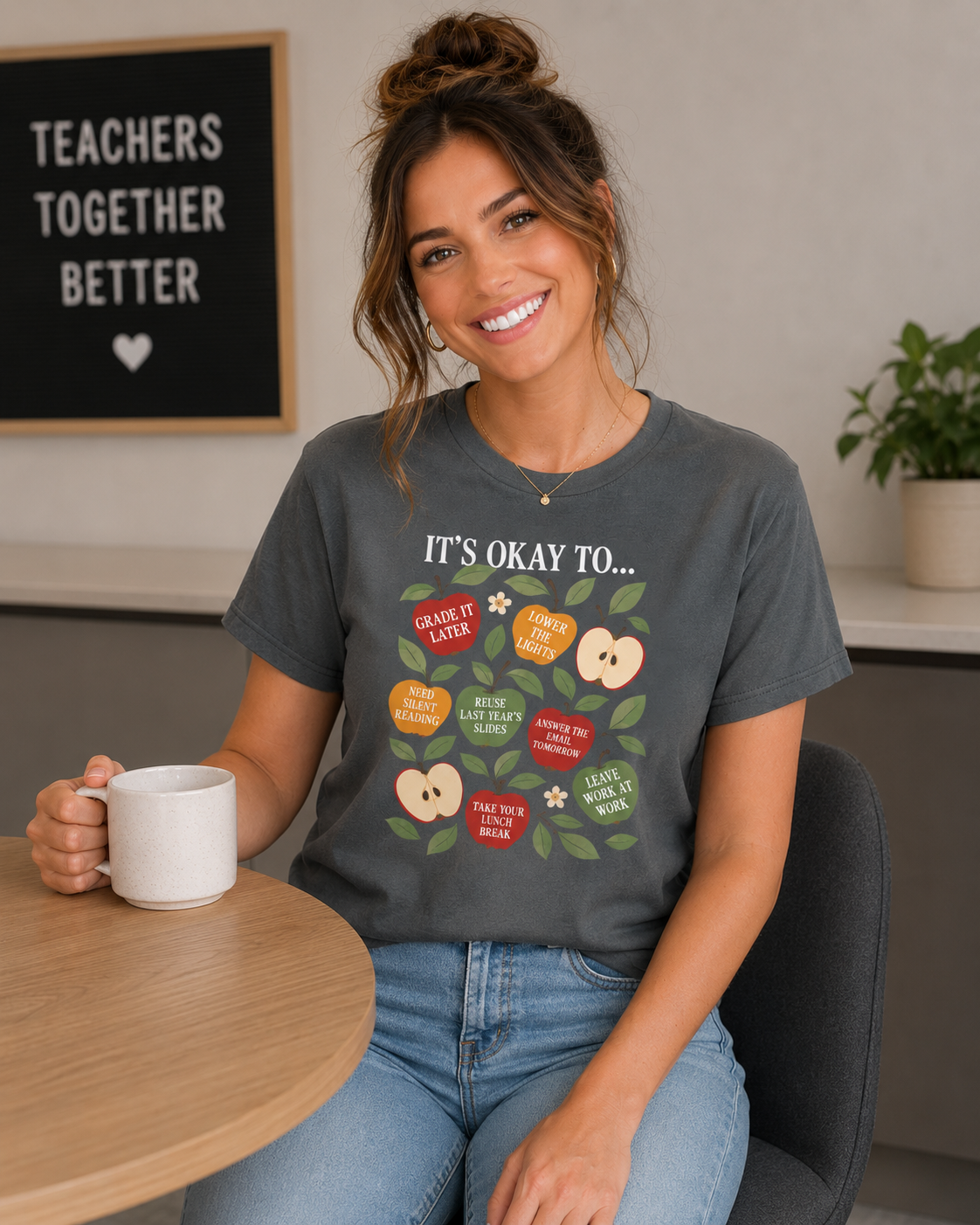 Woman wearing a Comfort Colors 1717 t-shirt in pepper gray featuring colorful apple illustrations with teacher self care sayings including “Grade It Later,” “Reuse Last Year’s Slides,” and “Leave Work at Work” while sitting with a coffee mug.