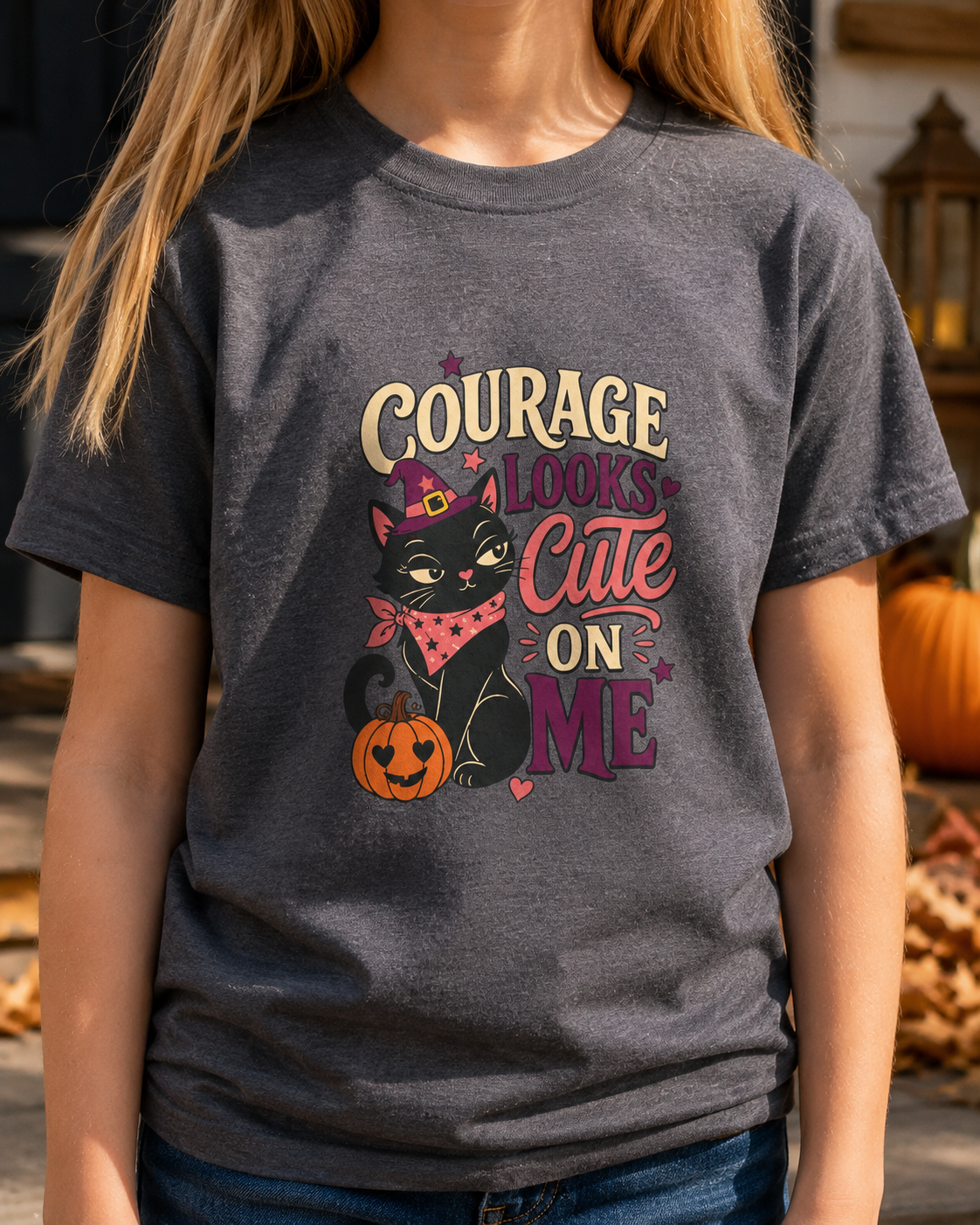 Girl wearing a deep heather grey color youth T-shirt featuring a witchy black cat, heart-eyed jack-o’-lantern, colorful stars, and the phrase “Courage Looks Cute on Me.”