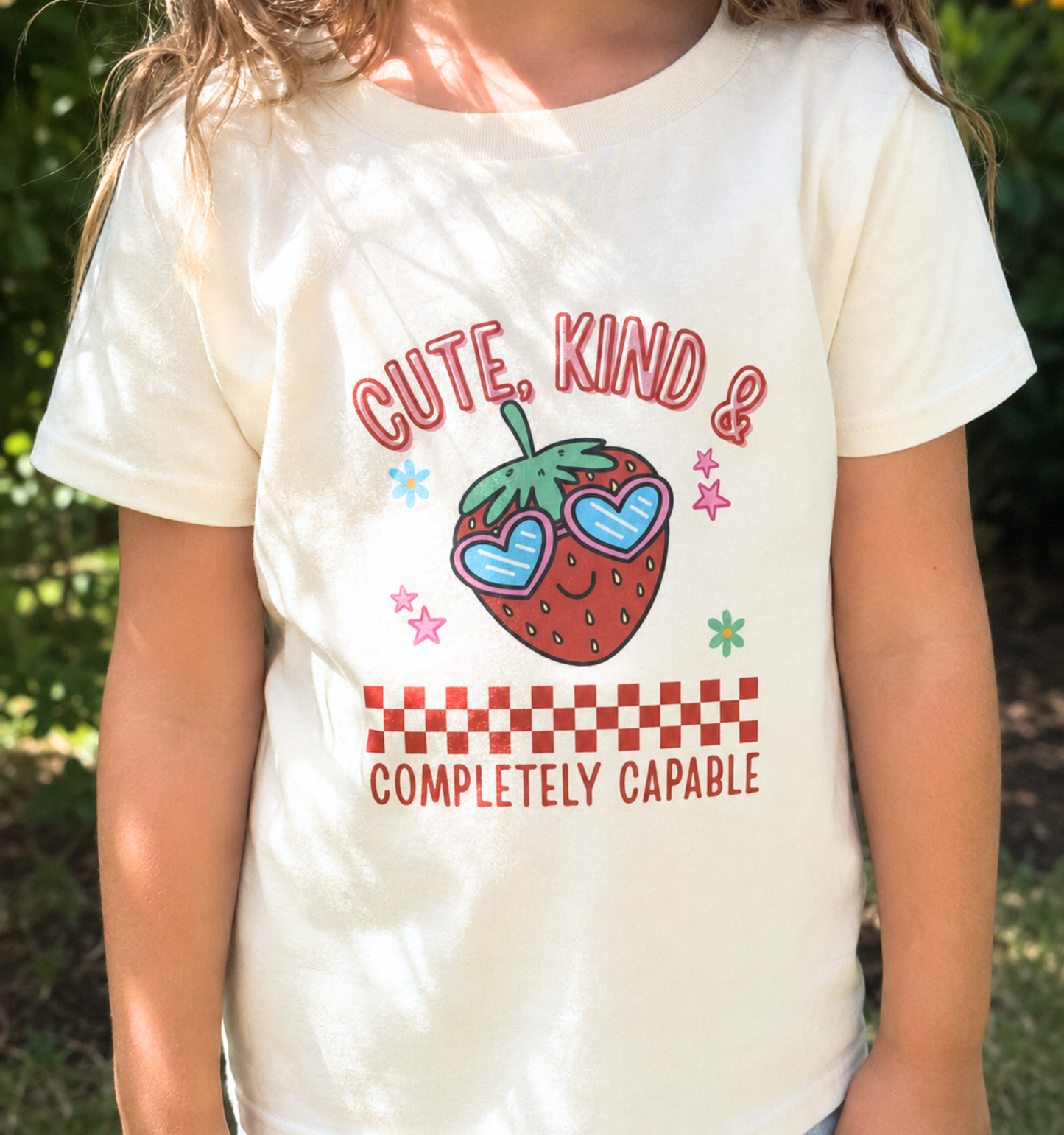 Child wearing a natural toddler T-shirt featuring a smiling strawberry in heart-shaped sunglasses and the phrase “Cute, Kind & Completely Capable.”