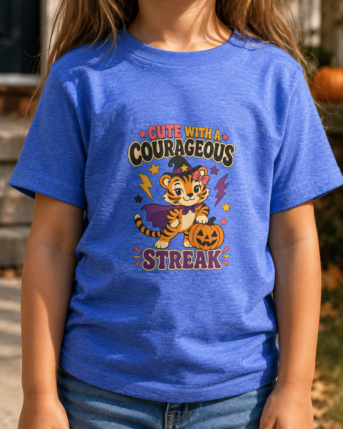 Child wearing a heather Columbia blue toddler T-shirt featuring a witchy tiger, jack-o’-lantern, colorful stars, and the phrase “Cute With a Courageous Streak.”