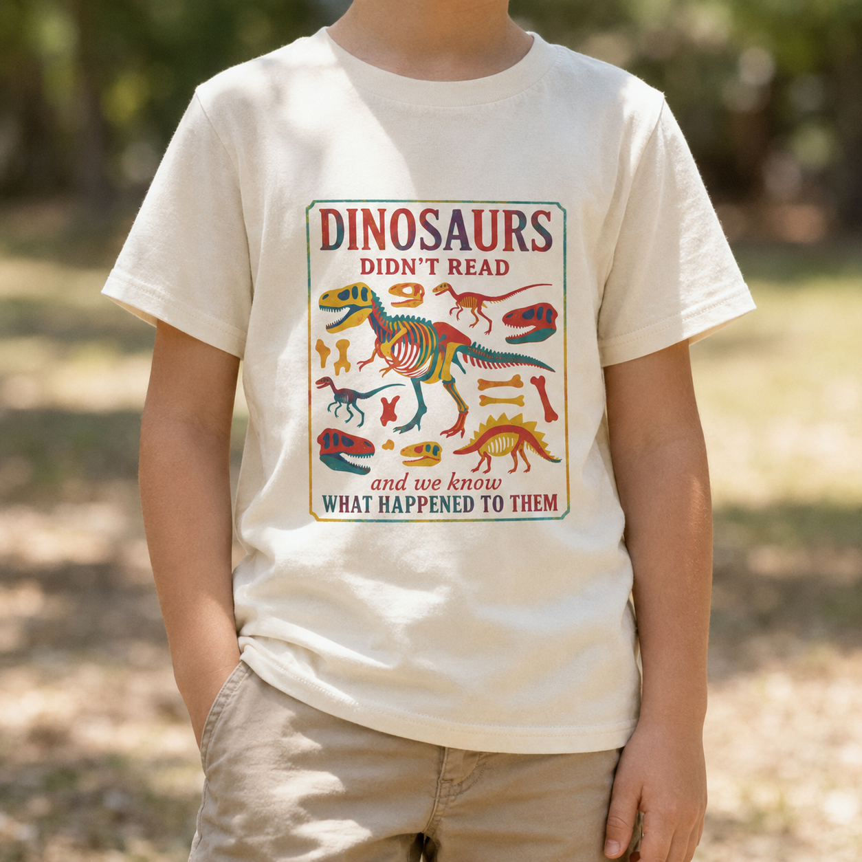 youth t-shirt in Natural featuring a colorful dinosaur skeleton graphic with the humorous phrase "Dinosaurs Didn't Read and We Know What Happened to Them." Fun youth graphic tee for young readers, book lovers, school, library visits, and back-to-school outfits.