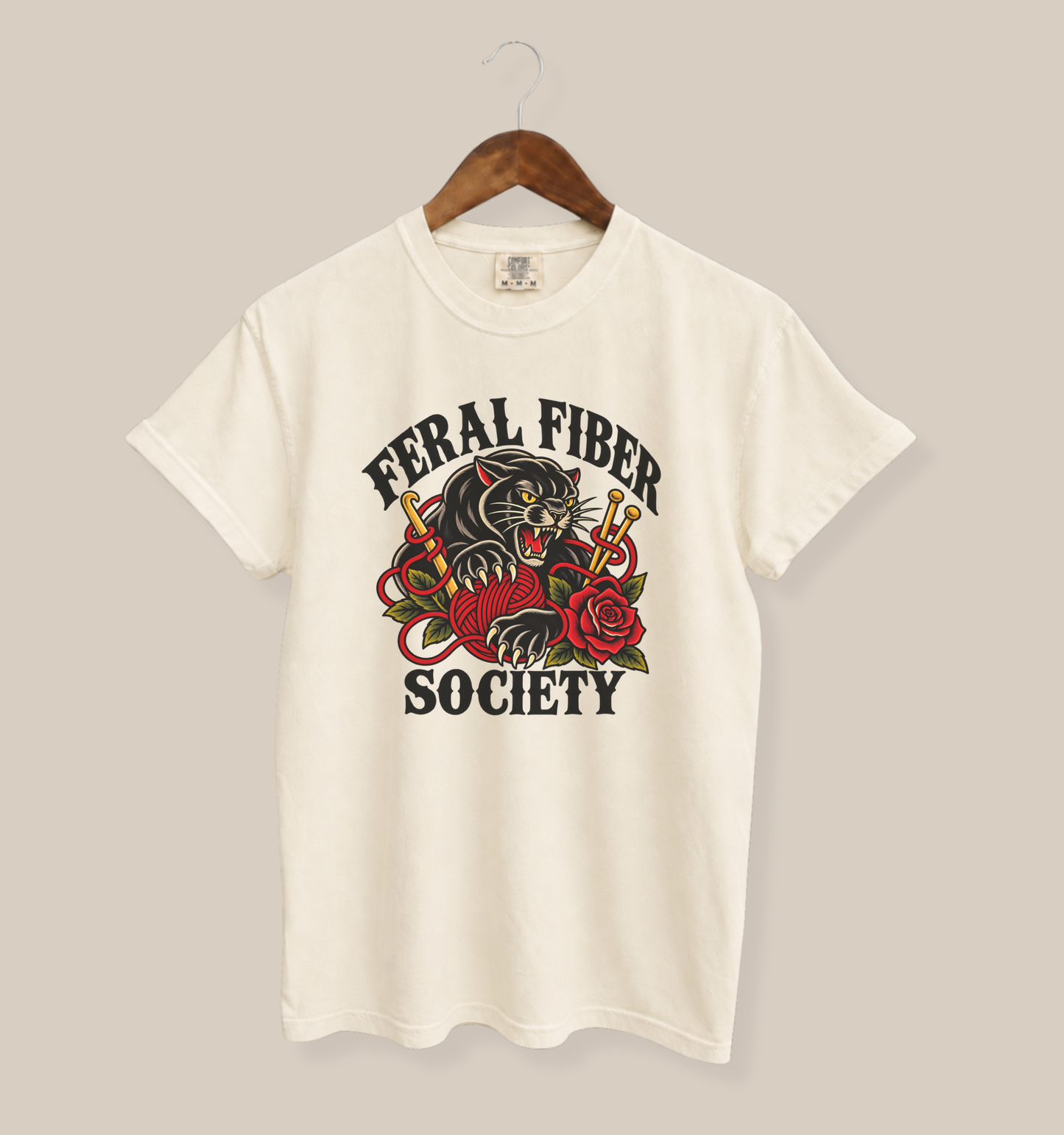 An off-white short-sleeve t-shirt hanging on a wooden hanger against a solid beige background. The shirt features a graphic print on the front with the text 'FERAL FIBER SOCIETY' surrounding an illustration of a black panther, red roses, and gold chains.