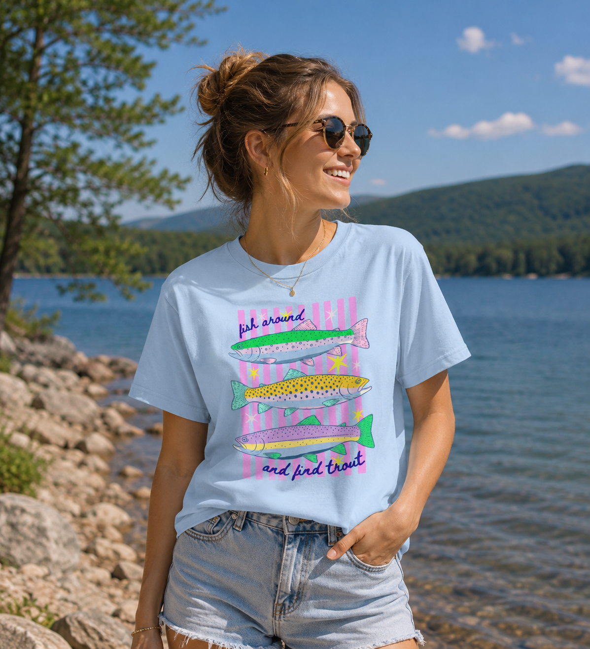 Woman wearing a light blue Bella + Canvas 3001 t-shirt featuring a colorful “Fish Around and Find Trout” graphic with illustrated trout and pink striped accents beside a lake.
