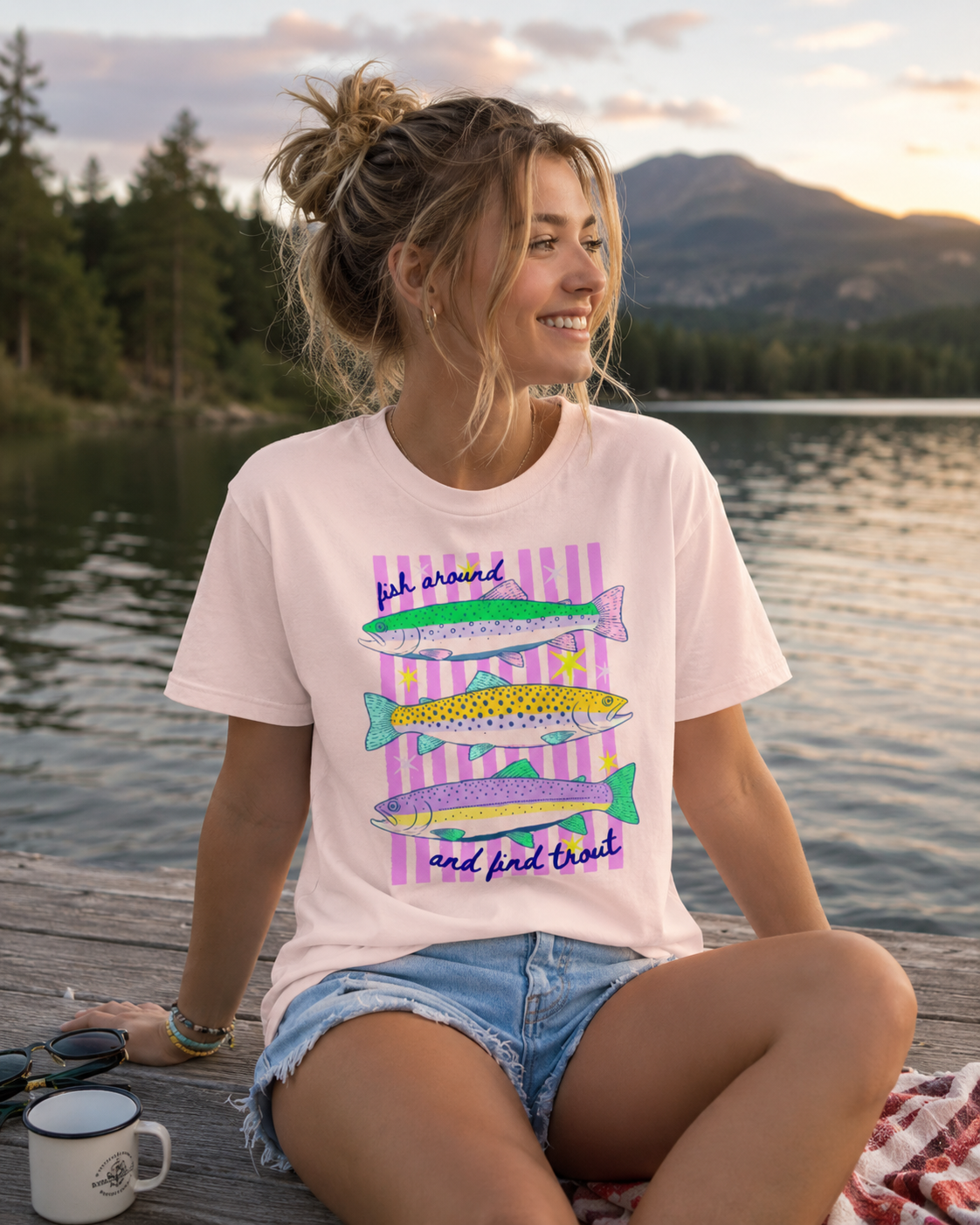 Woman wearing a soft pink Bella + Canvas 3001 t-shirt featuring a colorful “Fish Around and Find Trout” graphic while sitting on a lakeside dock at sunset.