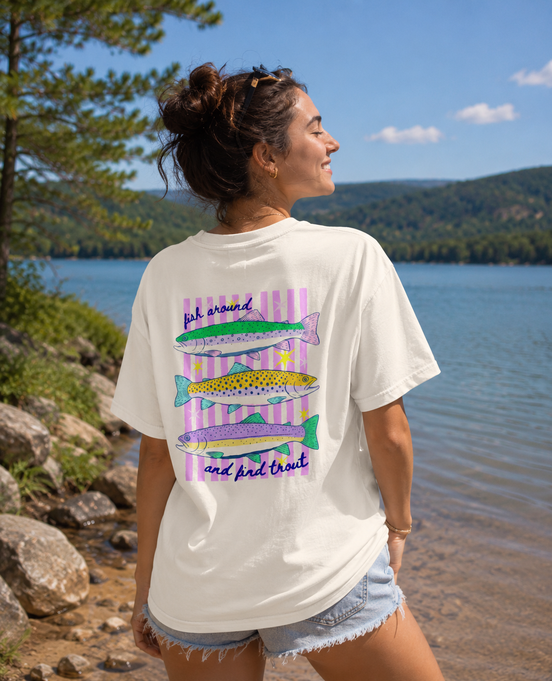Woman standing by a lake wearing an ivory Comfort Colors 1717 t-shirt with a colorful “Fish Around and Find Trout” back graphic featuring illustrated trout and pink striped accents.