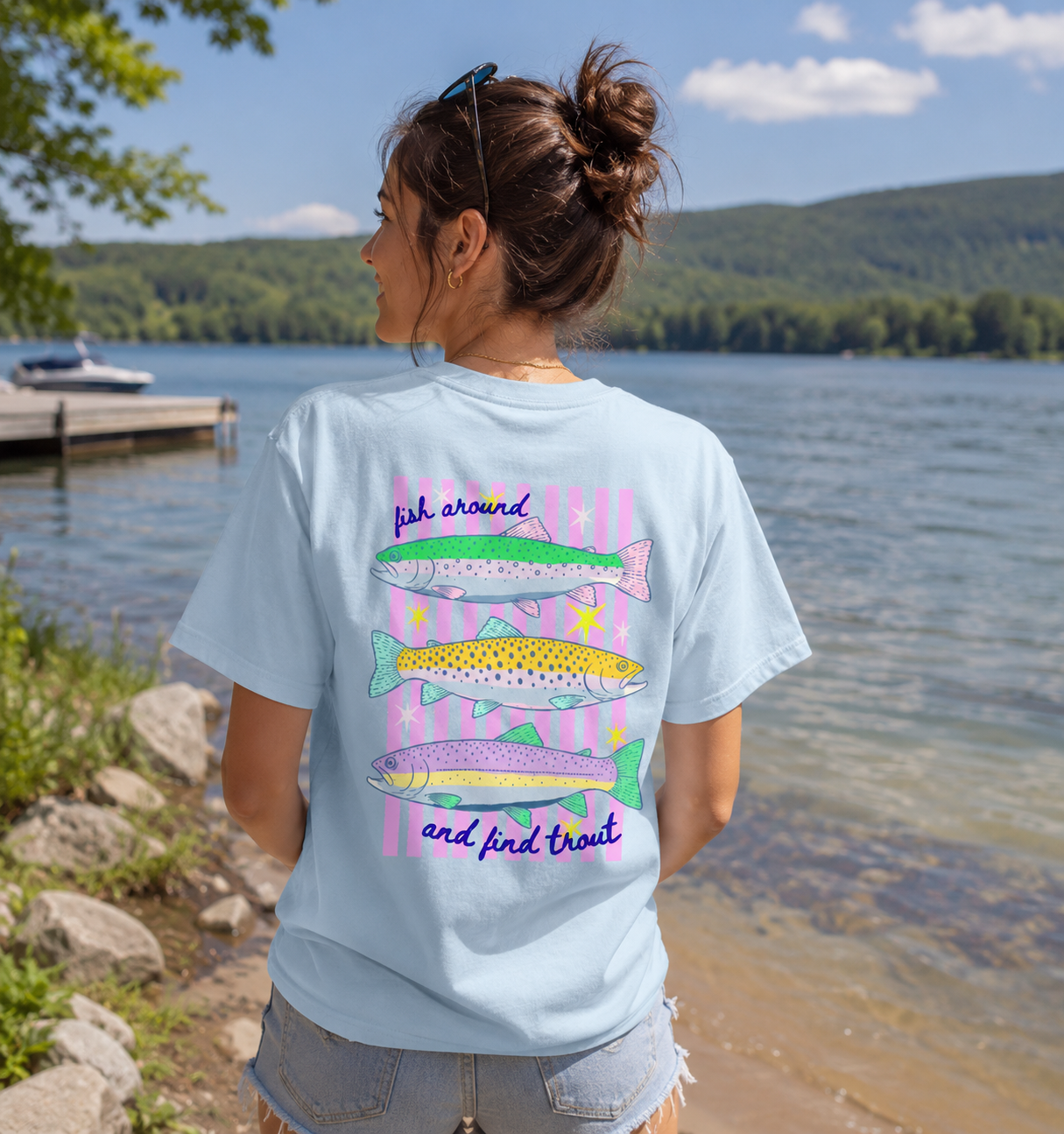 Woman standing lakeside wearing a chambray blue Comfort Colors 1717 t-shirt with a colorful “Fish Around and Find Trout” back graphic featuring illustrated trout and pink striped accents.