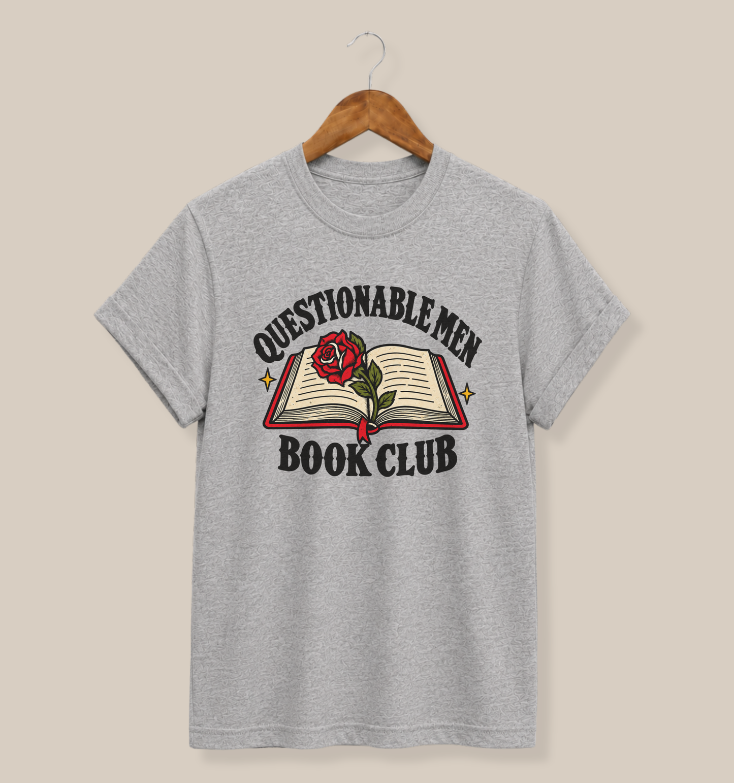 A heather gray short-sleeve t-shirt hanging on a wooden hanger against a solid beige background. The shirt features a graphic print of an open book with a red rose and the text 'QUESTIONABLE MEN BOOK CLUB'.