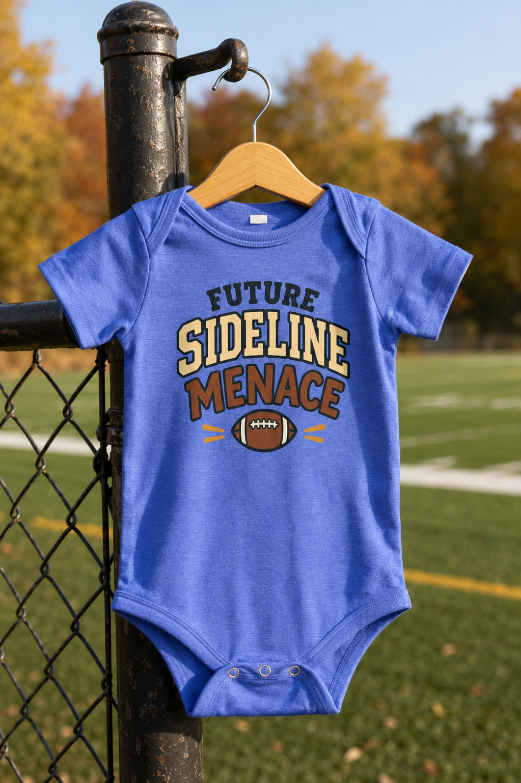 Heather Columbia blue baby one piece featuring a football graphic and the playful phrase “Future Sideline Menace,” hanging beside a football field.