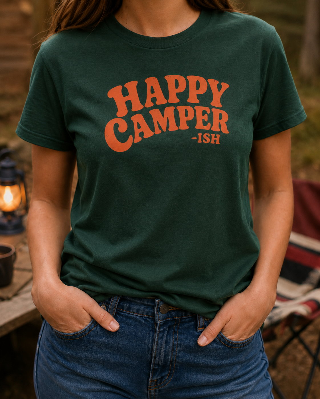 Woman wearing a forest green adult T-shirt featuring distressed orange retro lettering and the humorous phrase “Happy Camper-ish.”