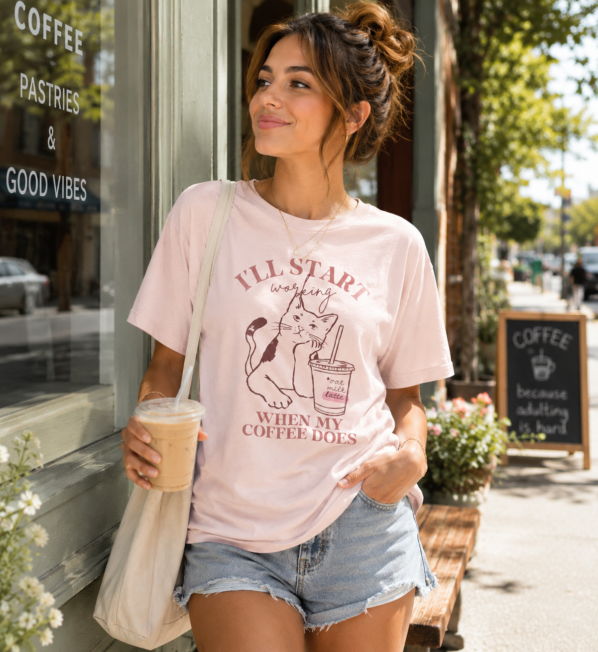 Woman wearing a soft pink Bella + Canvas 3001 t-shirt featuring an “I’ll Start Working When My Coffee Does” graphic with a cat and iced coffee illustration outside a coffee shop.