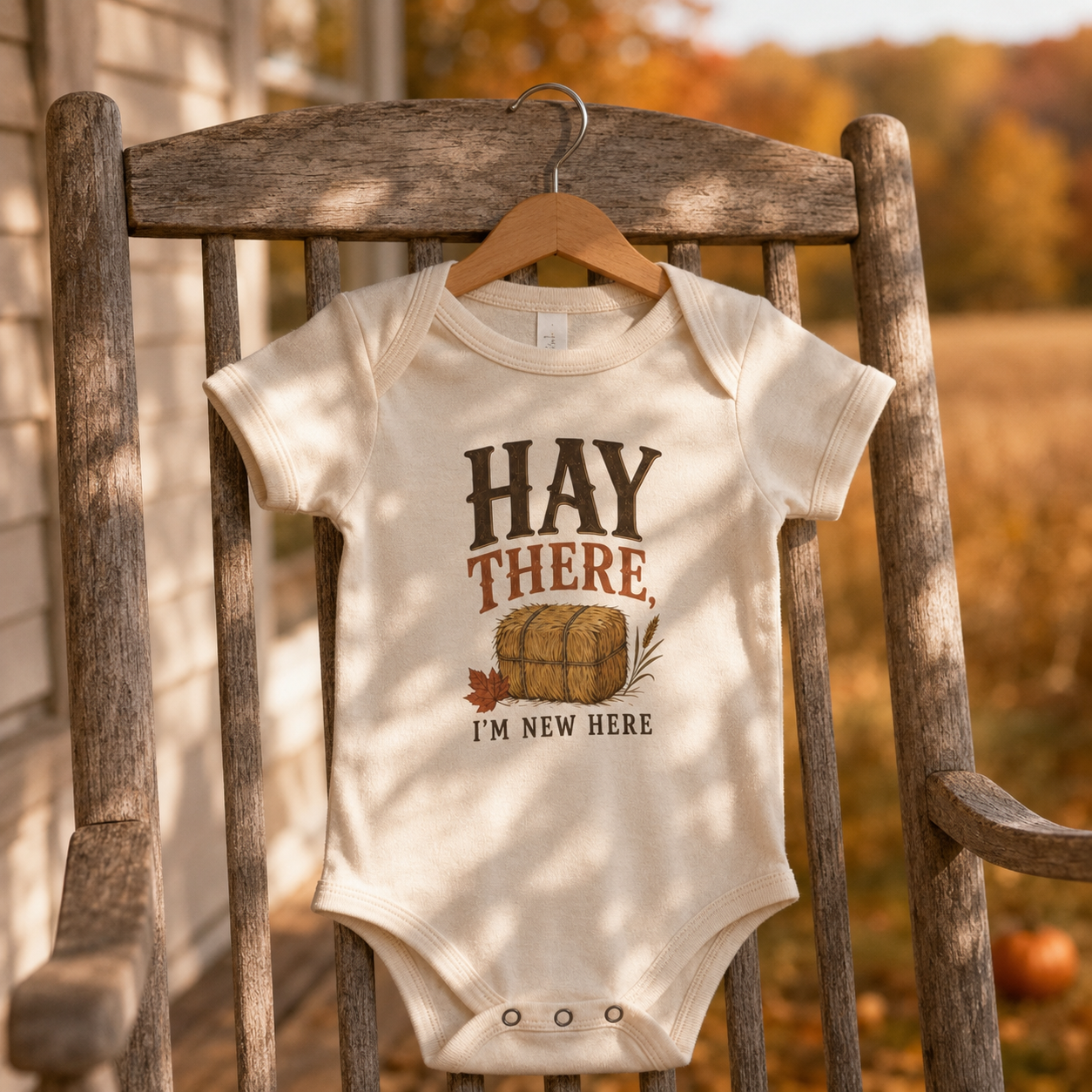 Natural Bella + Canvas 100B infant one piece featuring the phrase "Hay There, I'm New Here" with a hay bale and autumn leaves illustration. Cute fall baby bodysuit for newborns and infants, perfect for baby's first autumn, pumpkin patch visits, hayrides, baby showers, fall photos, and seasonal celebrations.