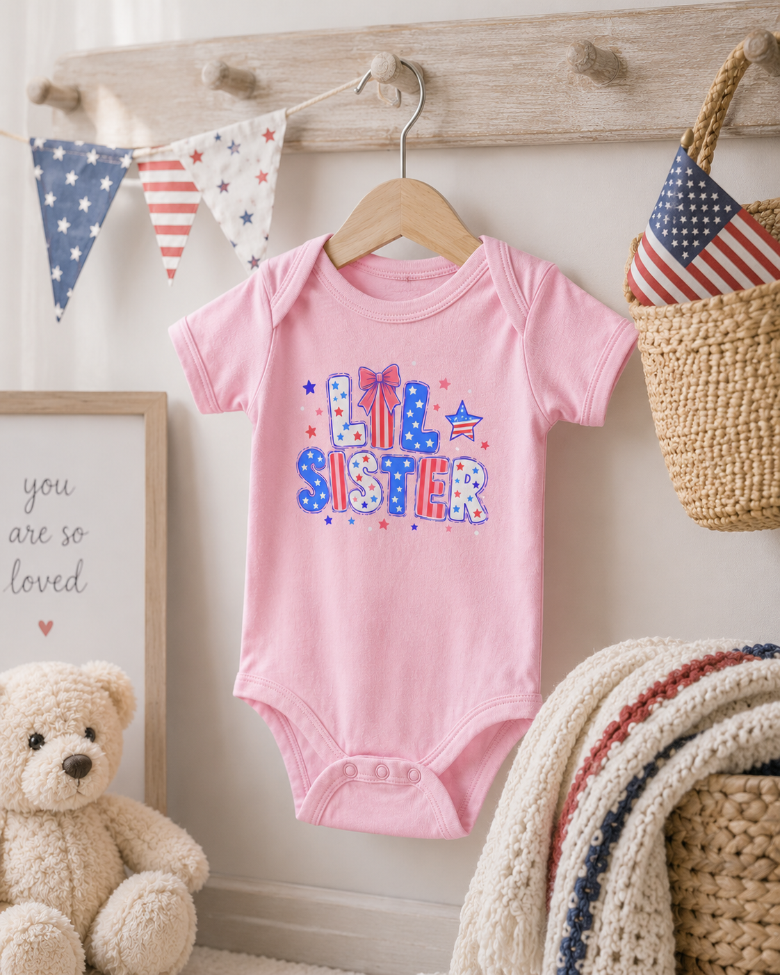 Light pink Bella + Canvas 100B infant one piece with patriotic “Lil Sister” graphic displayed in a cozy nursery setting with American flag decor, soft blankets, and Fourth of July themed accents.