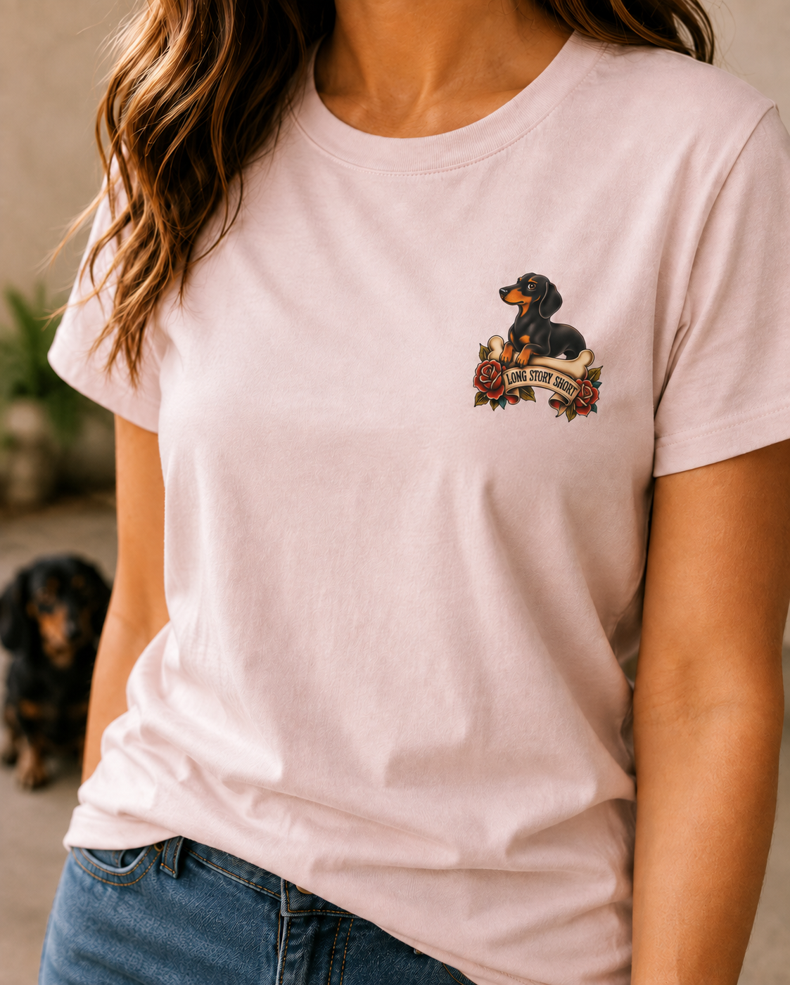 A woman wearing a light pink crewneck t-shirt with a small graphic of a dachshund on the left chest, paired with blue jeans.