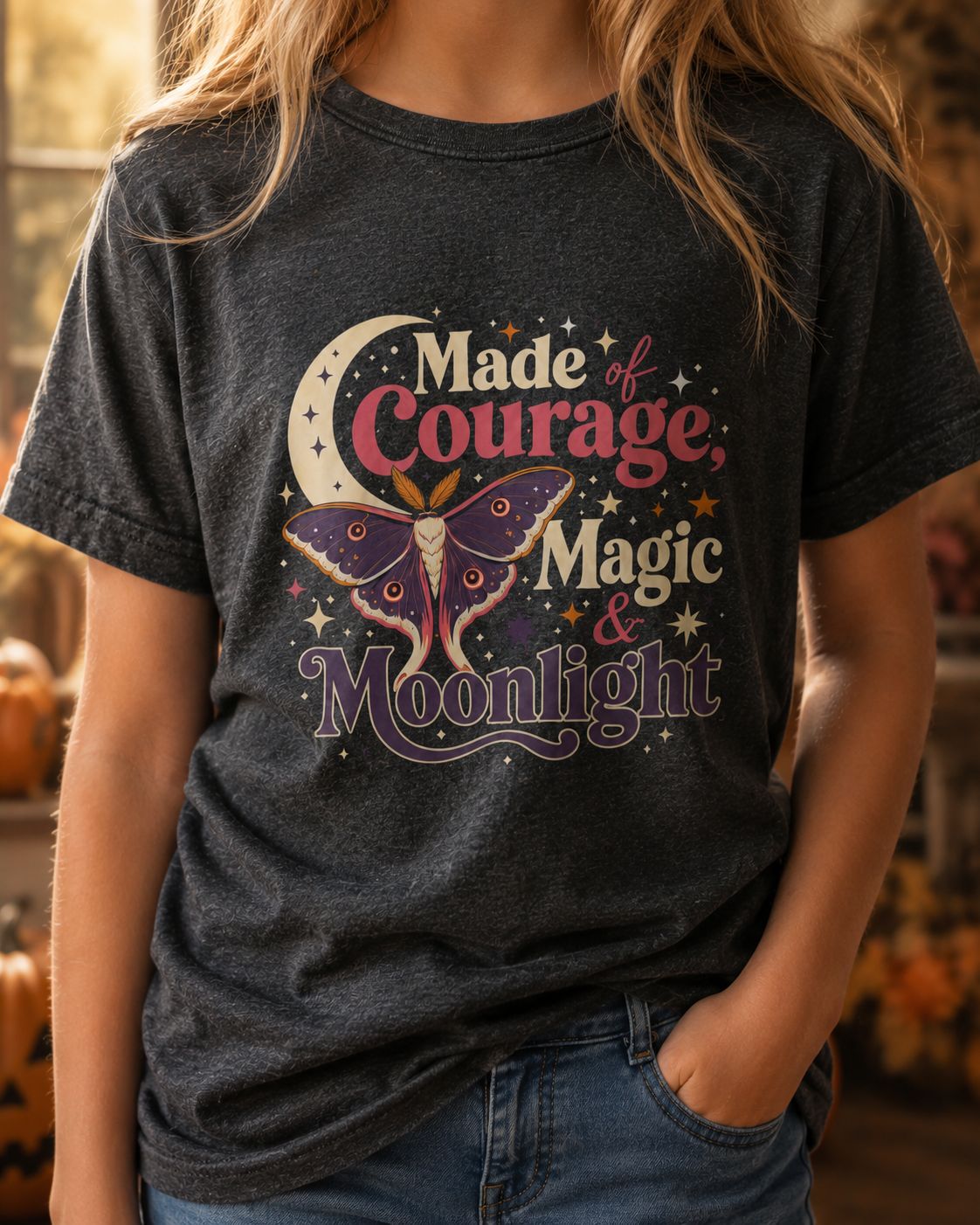Girl wearing a dark grey heather youth T-shirt featuring a colorful celestial moth, crescent moon, sparkling stars, and the phrase “Made of Courage, Magic & Moonlight.”