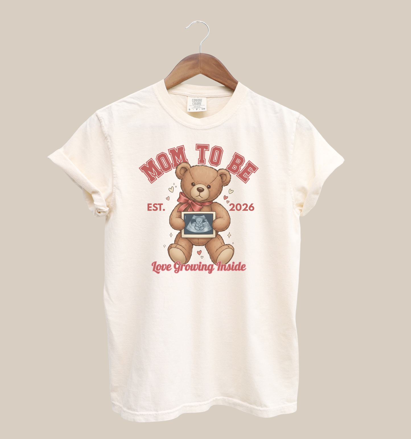 White t-shirt with teddy bear graphic and text 'Mom to Be' on a beige background