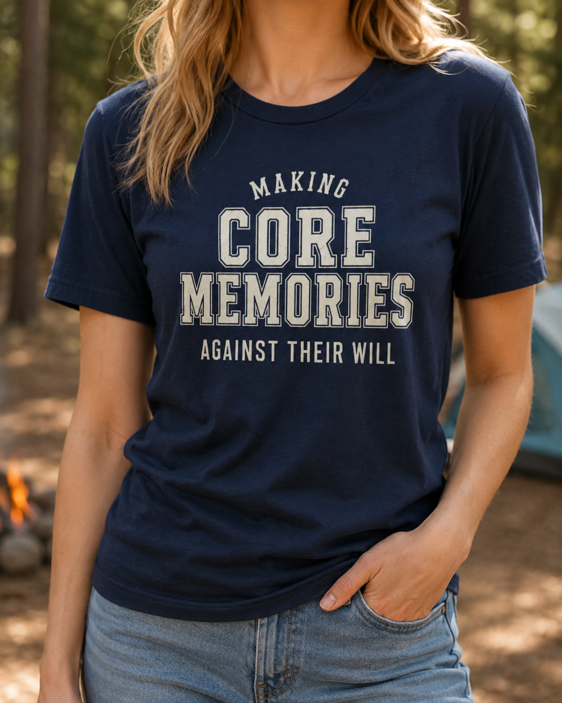 Woman wearing a navy adult T-shirt featuring distressed cream varsity lettering and the humorous phrase “Making Core Memories Against Their Will” at a campsite.