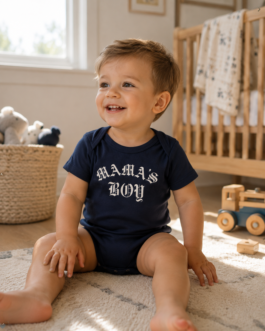 Baby wearing a navy Bella + Canvas 100B infant one piece featuring a white “Mama’s Boy” gothic-style graphic while sitting in a cozy neutral nursery with warm natural lighting.