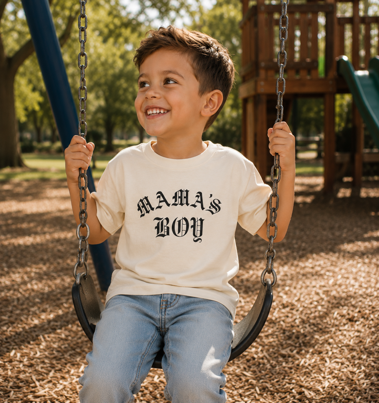 Natural Bella + Canvas toddler tee featuring a bold black “Mama’s Boy” gothic-style graphic, modeled on a young boy at a playground swing set for trendy everyday kids streetwear.