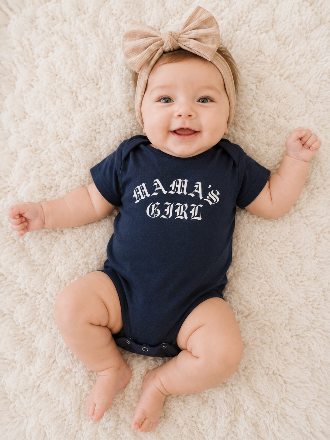 Baby girl wearing a navy Bella + Canvas 100B infant one piece featuring a white “Mama’s Girl” gothic-style graphic while lying on a soft cream blanket with a neutral bow headband.