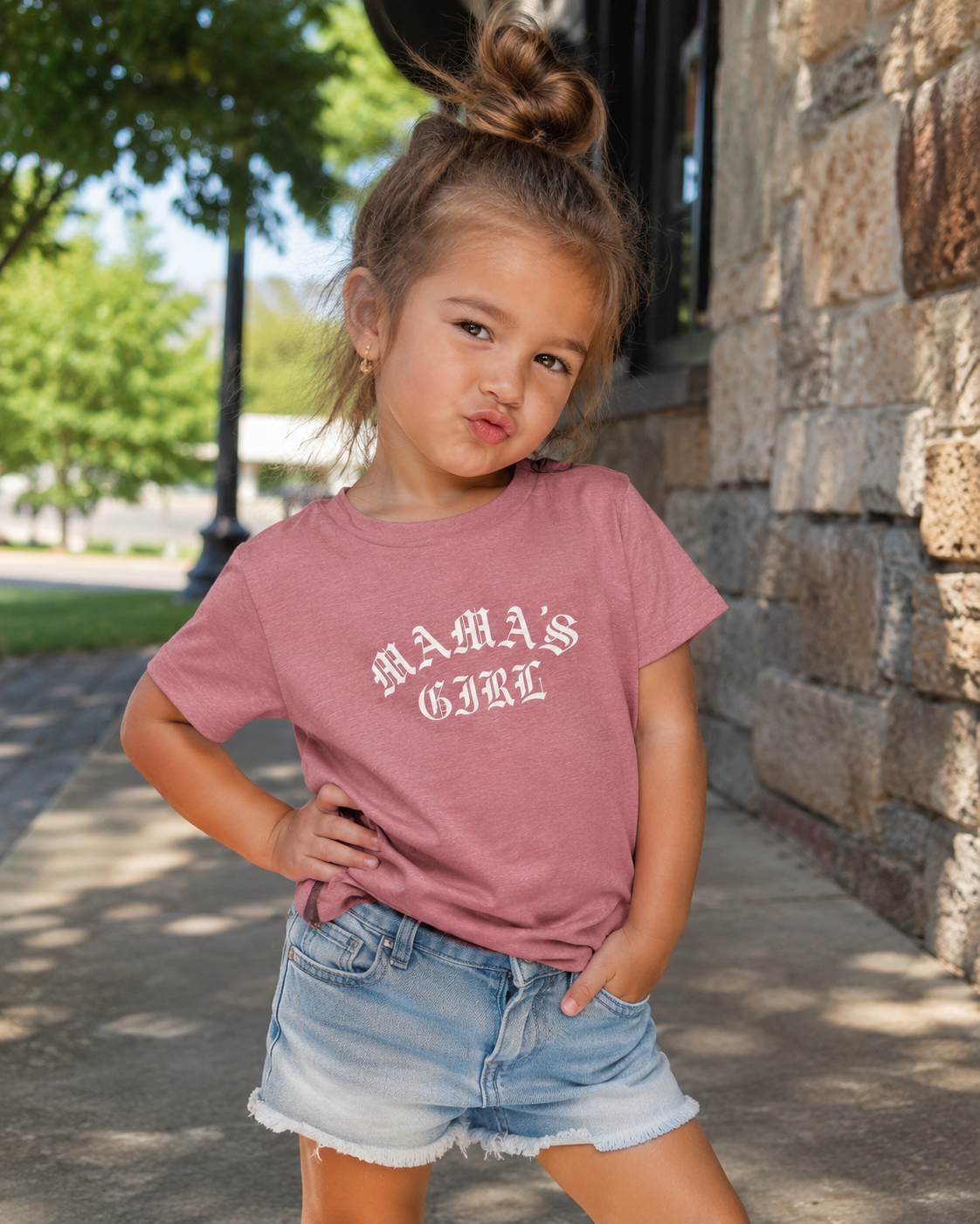 Toddler girl wearing a heather mauve Bella + Canvas toddler tee with a cream “Mama’s Girl” gothic-style graphic, paired with denim shorts in an outdoor lifestyle mockup for trendy mommy-and-me apparel.
