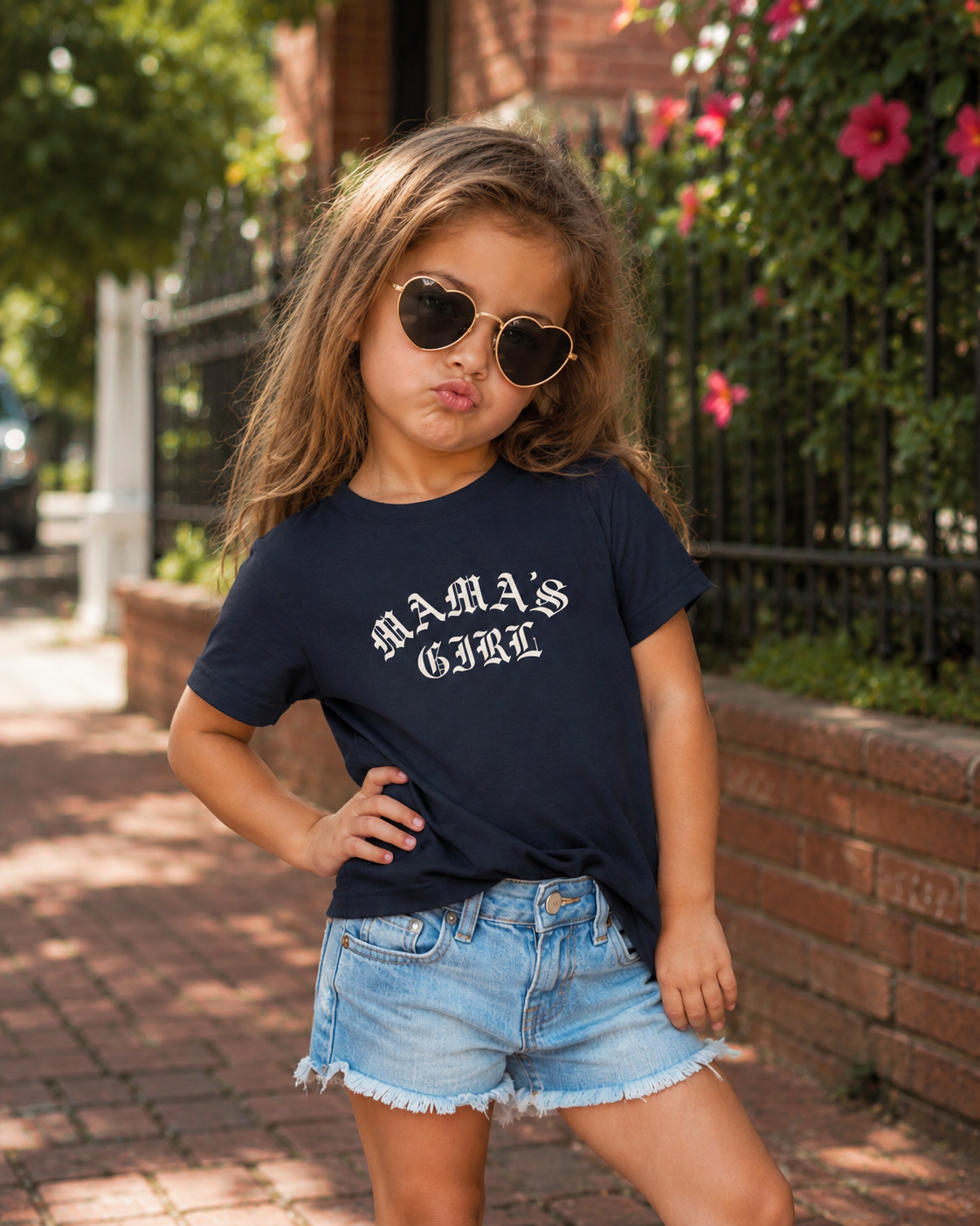 Toddler girl wearing a navy Bella + Canvas toddler tee with a white “Mama’s Girl” gothic-style graphic, styled with denim shorts and sunglasses in a playful outdoor lifestyle mockup.