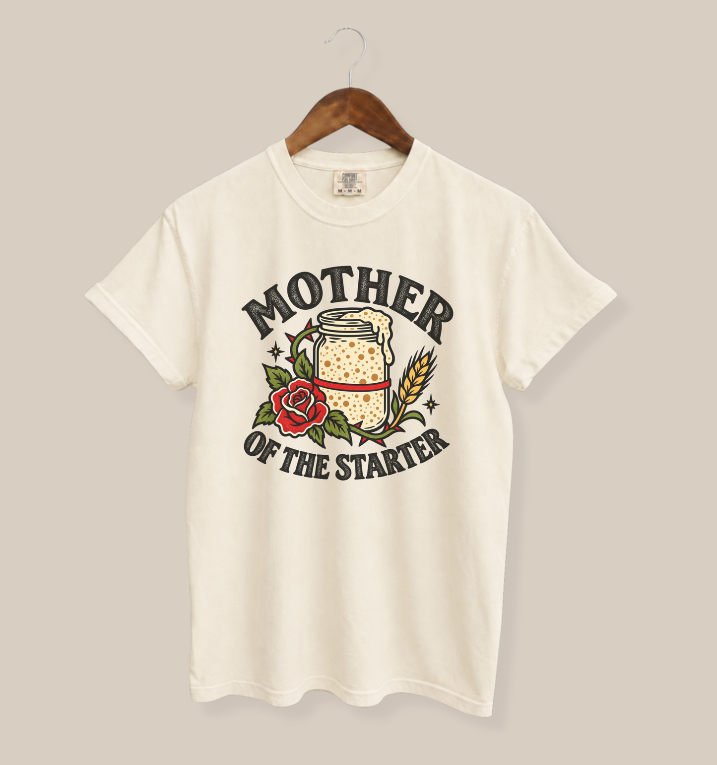 An ivory short-sleeve t-shirt hanging on a wooden hanger against a solid beige background. The shirt features a graphic print with the text 'MOTHER OF THE STARTER' in black, surrounding an illustration of a glass jar filled with a dotted yellow liquid, a red rose, and wheat stalks.