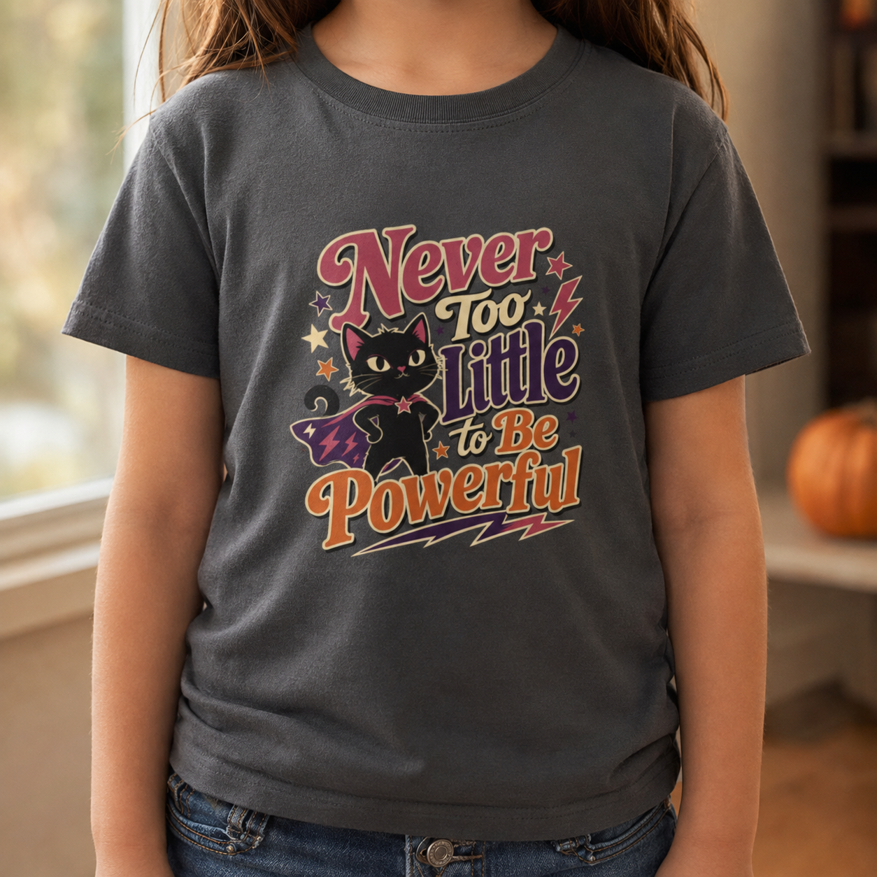 Girl wearing an asphalt grey toddler T-shirt featuring a superhero black cat in a purple cape, colorful stars, lightning bolts, and the phrase “Never Too Little to Be Powerful.”