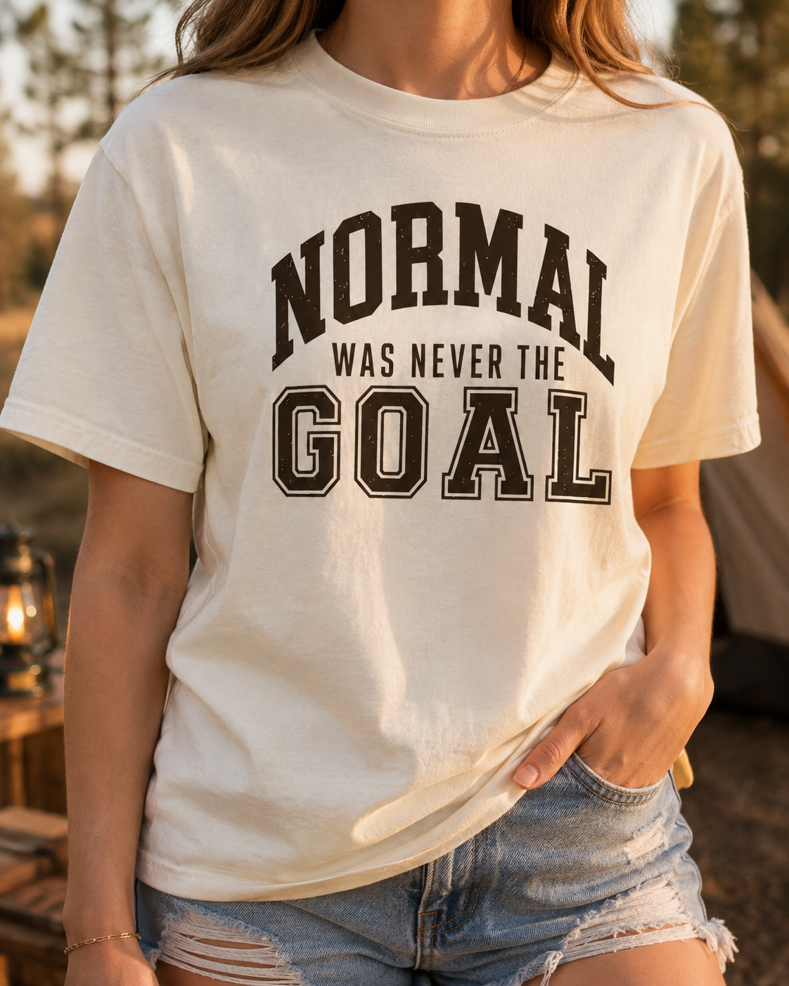Woman wearing a natural adult T-shirt featuring distressed brown varsity lettering and the humorous phrase “Normal Was Never the Goal.”