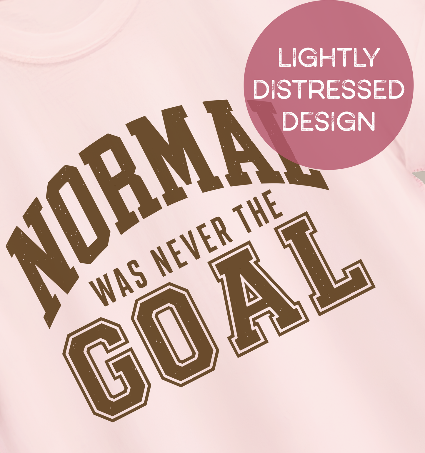 Close-up of a soft pink adult T-shirt featuring lightly distressed brown varsity lettering and the humorous phrase “Normal Was Never the Goal.”