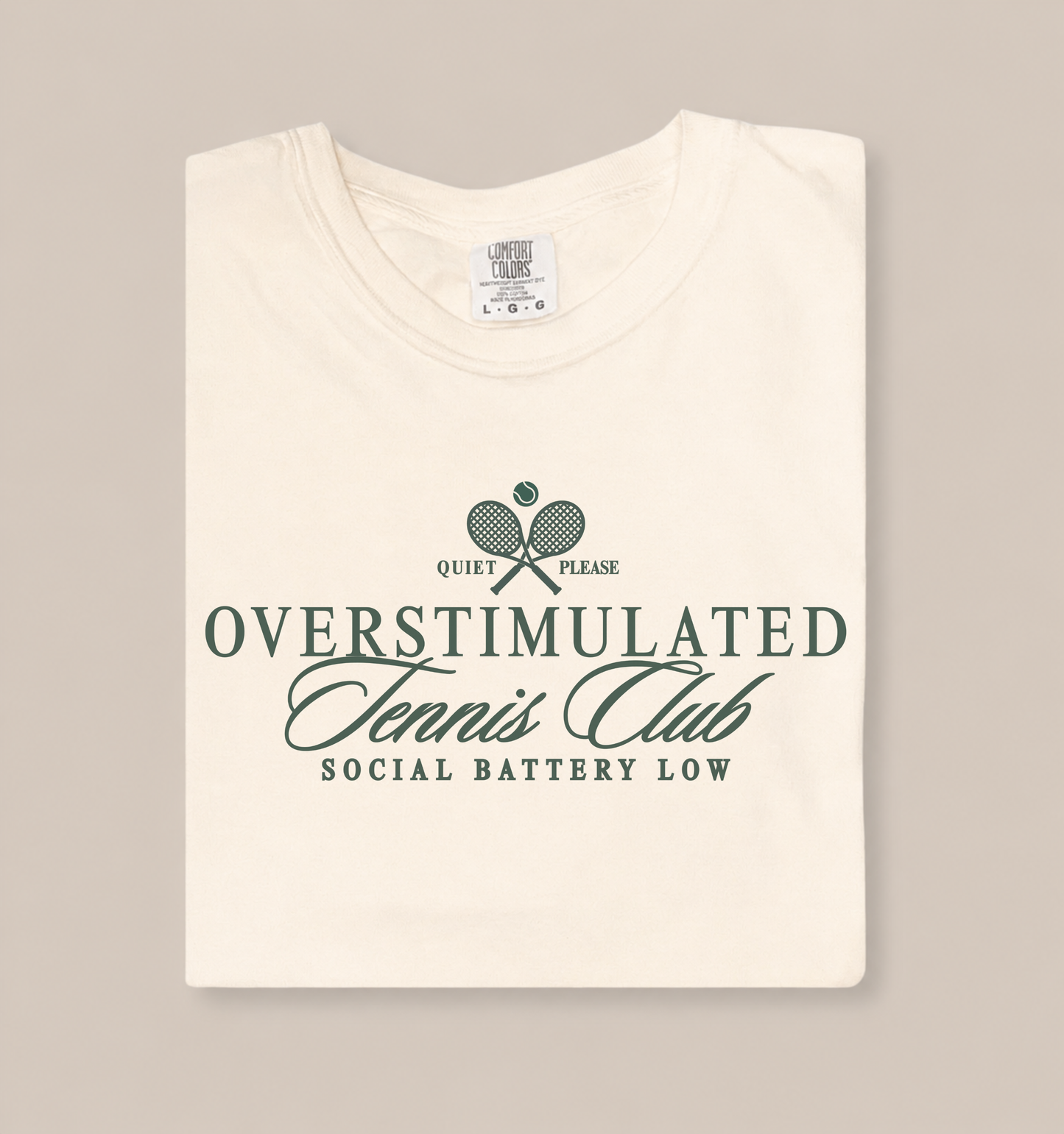 Folded ivory Comfort Colors 1717 t-shirt featuring a vintage-inspired “Overstimulated Tennis Club” graphic with crossed tennis rackets and the phrase “Social Battery Low” in dark green lettering.