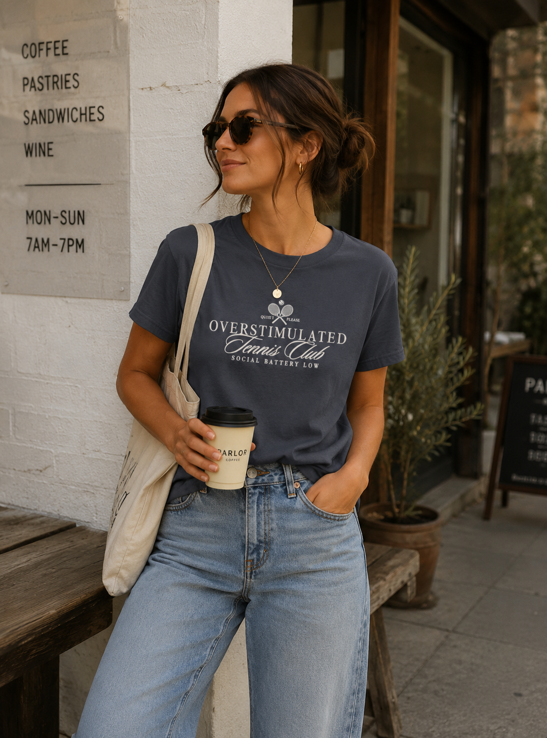 Woman wearing a navy Comfort Colors 1717 t-shirt featuring a vintage-inspired “Overstimulated Tennis Club” graphic with crossed tennis rackets and the phrase “Social Battery Low” while holding coffee outside a café.