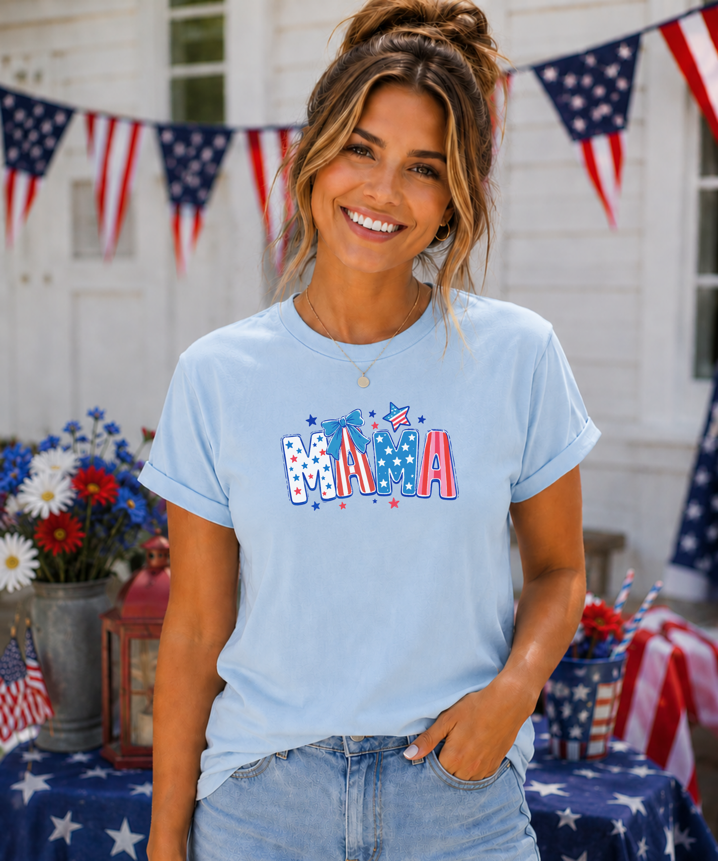 Woman wearing a baby blue Bella + Canvas t-shirt featuring a patriotic “MAMA” graphic with red, white, and blue lettering, stars, and a bow accent, styled in a festive Fourth of July setting with American flags and summer decor.