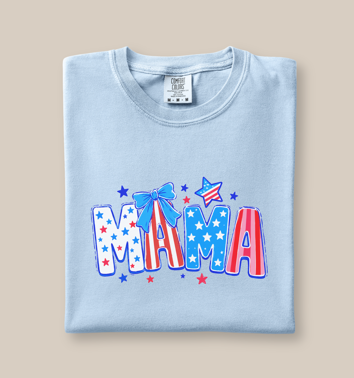 Adult Comfort Colors chambray blue t-shirt featuring a colorful patriotic “MAMA” graphic with red, white, and blue stars, striped lettering, and a blue bow accent in a playful Fourth of July style.