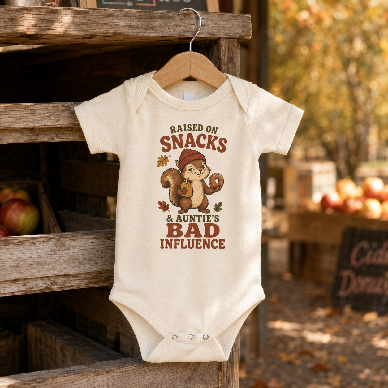 Natural baby one piece featuring a squirrel holding a donut and juice box with autumn leaves and the phrase “Raised on Snacks & Auntie’s Bad Influence,” hanging at a rustic apple orchard.