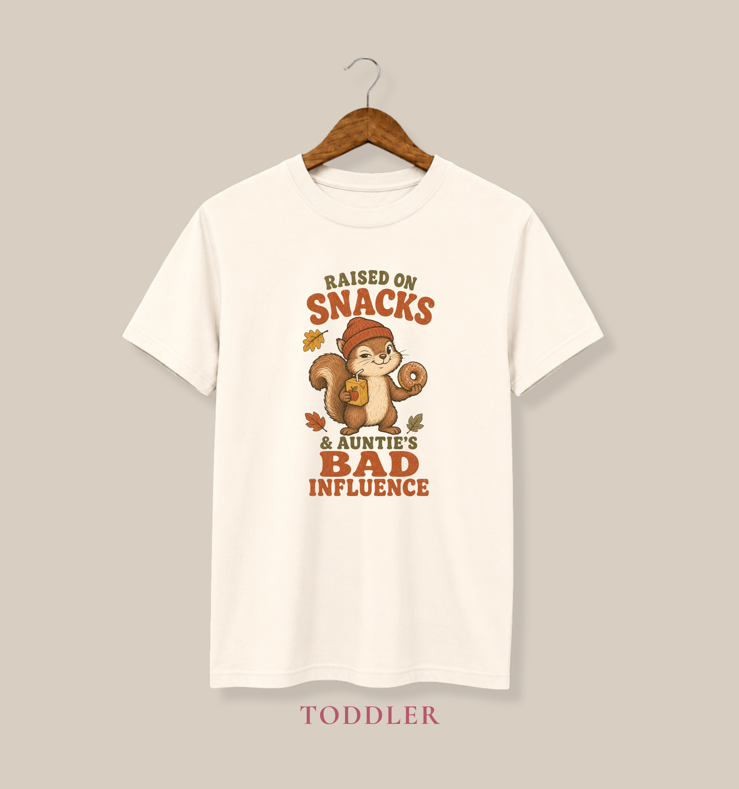Natural toddler T-shirt featuring a squirrel holding a donut and juice box, autumn leaves, and the phrase “Raised on Snacks & Auntie’s Bad Influence.”