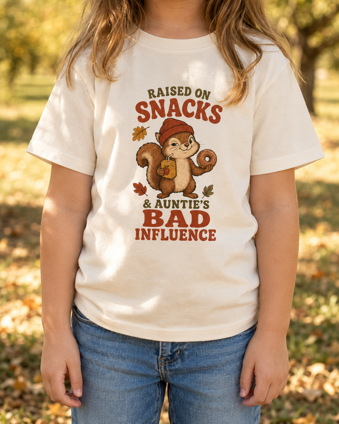 Child wearing a natural toddler T-shirt featuring a squirrel holding a donut and juice box, autumn leaves, and the phrase “Raised on Snacks & Auntie’s Bad Influence.”