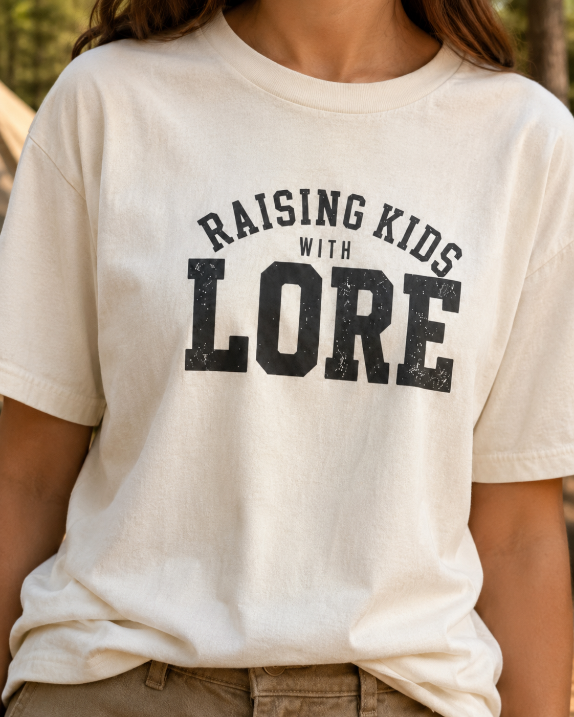 Woman wearing a natural BC3001 adult T-shirt featuring distressed charcoal varsity lettering and the humorous phrase “Raising Kids With Lore.”