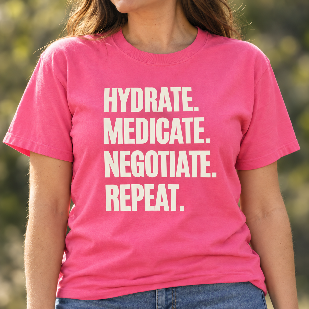 A woman wearing a bright pink crewneck t-shirt with the slogan "Hydrate. Medicate. Negotiate. Repeat."