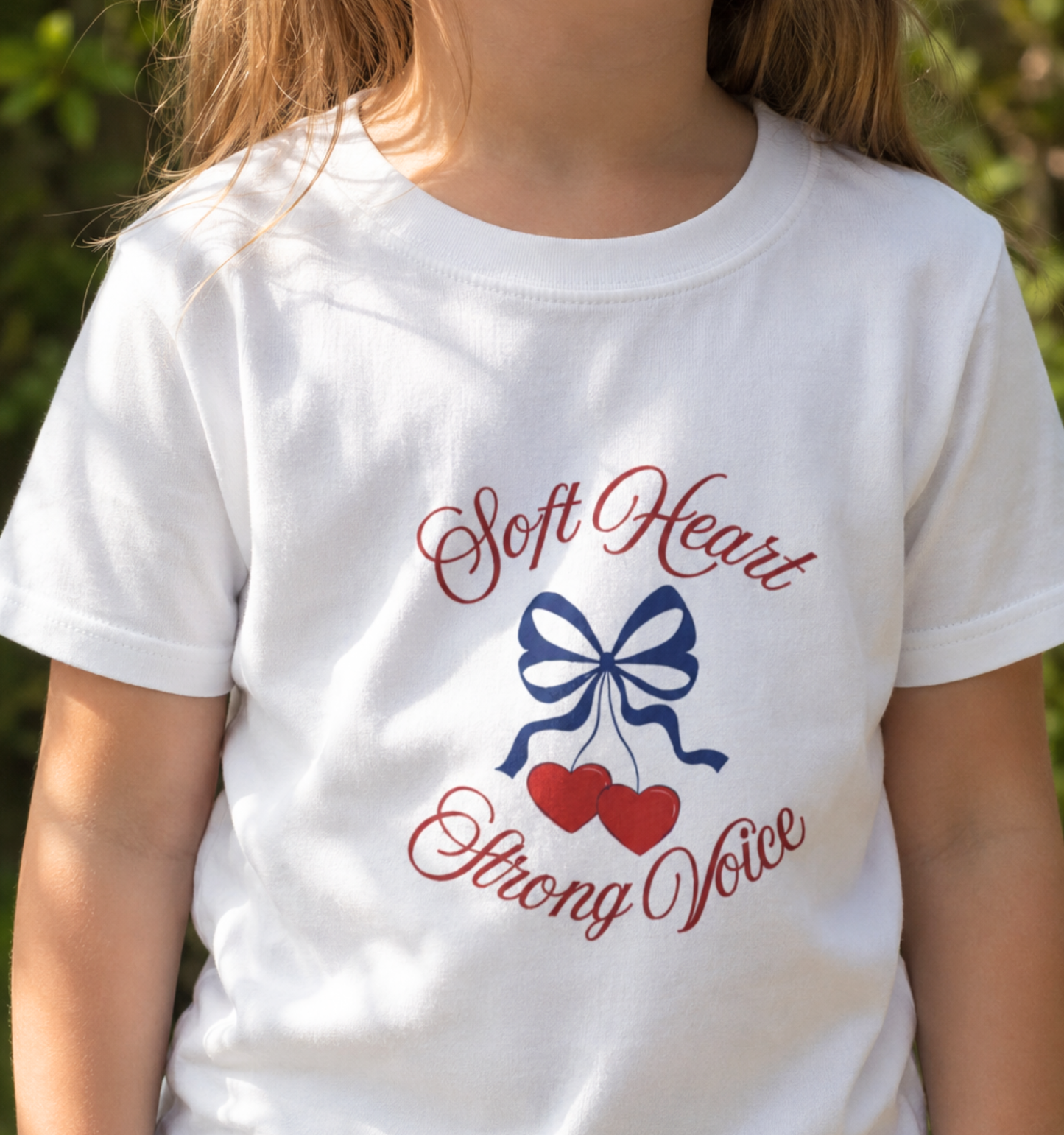 Child wearing a white toddler T-shirt featuring a navy bow, red hearts, and the empowering phrase “Soft Heart Strong Voice.”