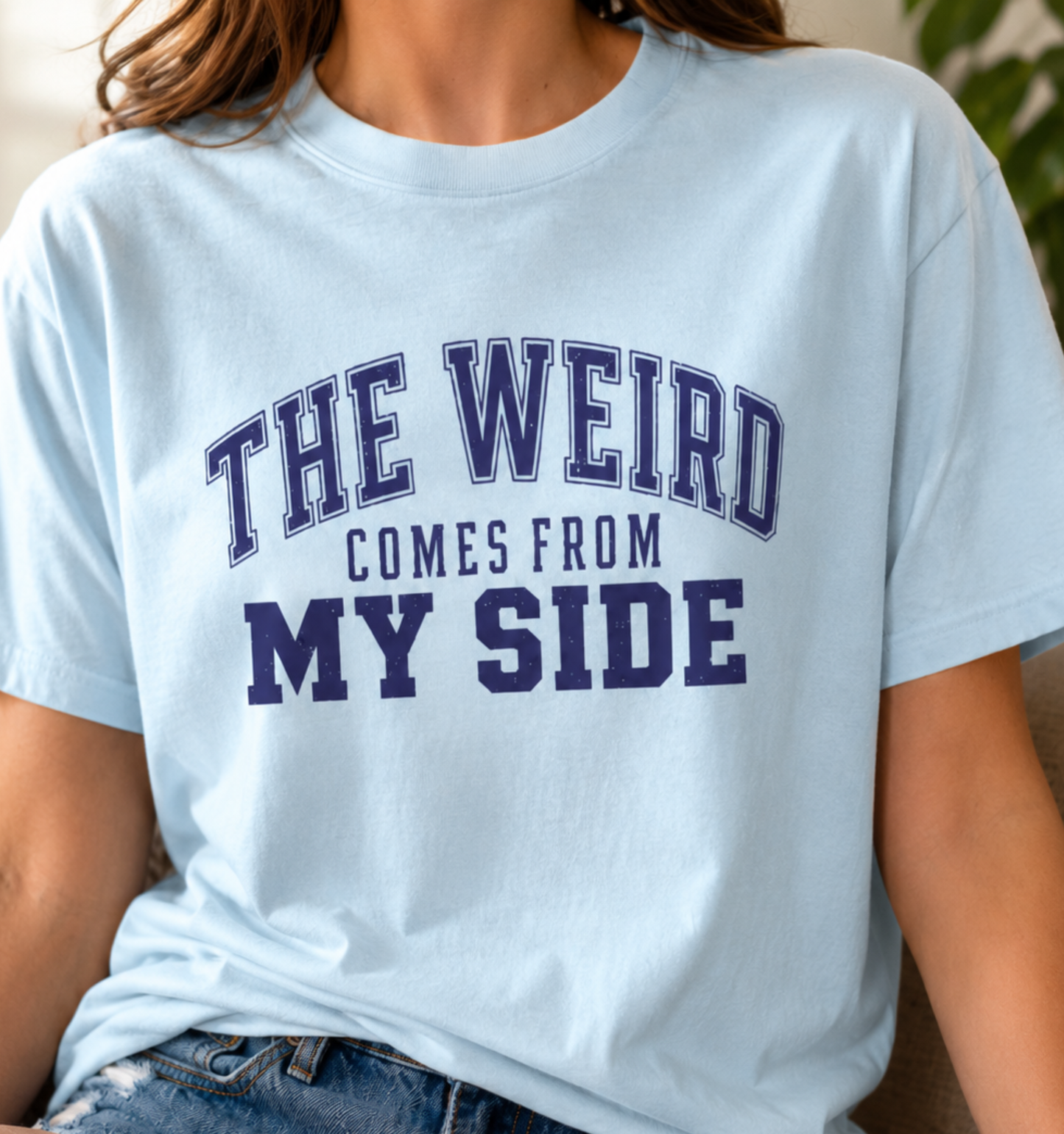 Woman wearing a baby blue adult T-shirt featuring a distressed navy varsity-style design with the humorous phrase “The Weird Comes From My Side.”