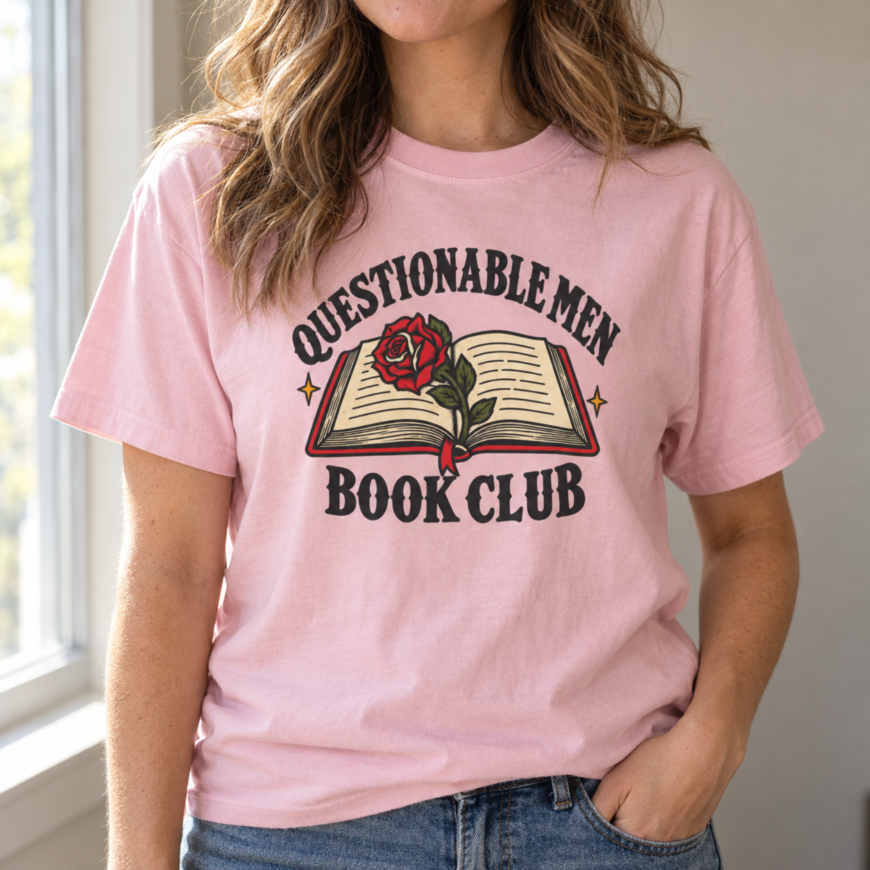 A woman wearing a light pink blossom graphic t-shirt with a 'QUESTIONABLE MEN BOOK CLUB' design featuring an open book and a red rose, paired with blue jeans.