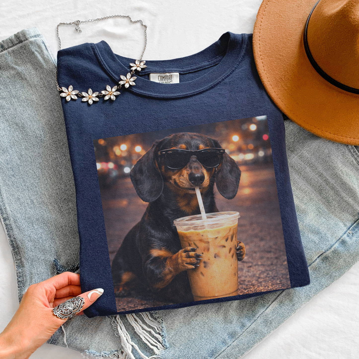 styled flat lay of navy blue graphic t-shirt featuring a dachshund dog in sunglasses drinking iced coffee with jeans hat and jewelry accessories