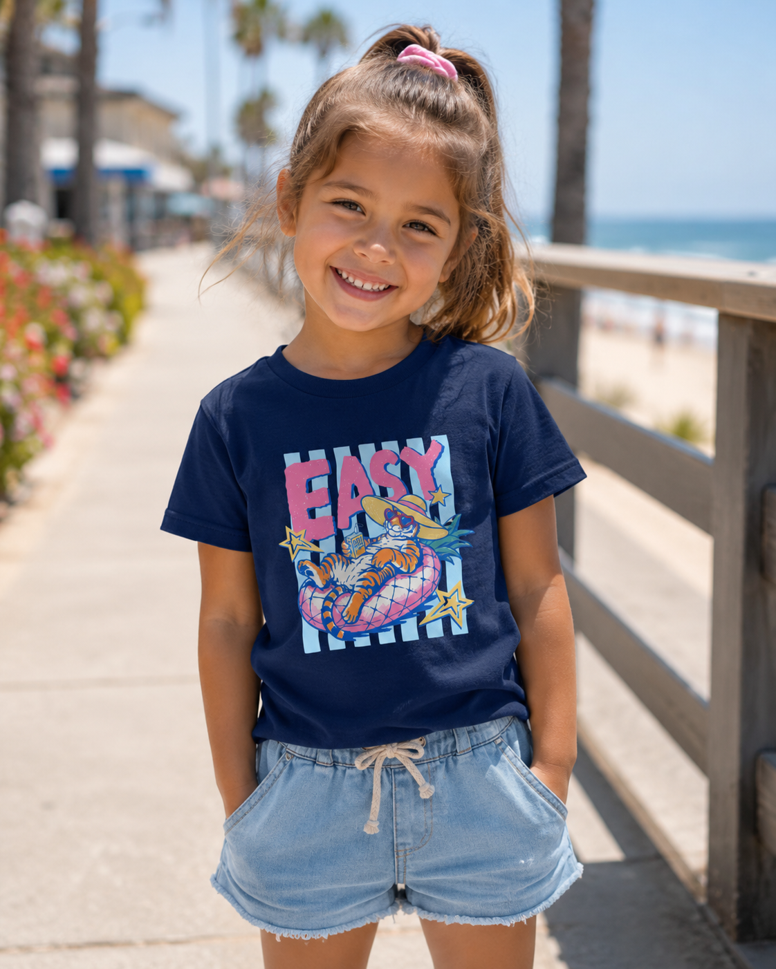cute toddler girl wearing navy blue easy tiger summer graphic tee with lounging tiger design while standing on beach boardwalk in sunshine