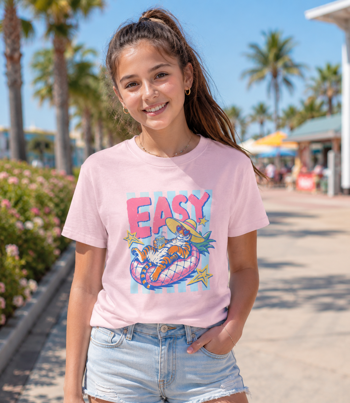 tween girl wearing pastel pink easy tiger summer graphic tee featuring lounging tiger pool float design on sunny tropical boardwalk